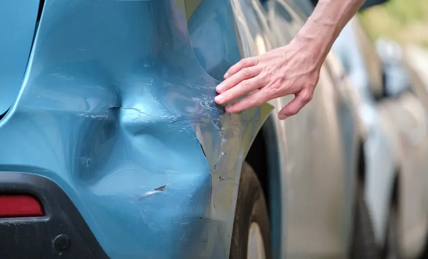 When Should You Hire a Lawyer After a Minor Fender Bender? A man's hand is examining his dented blue car bumper and damaged paint from a minor fender bender collision in Georgia.