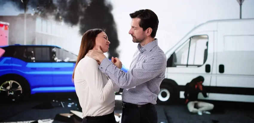 How Much Is Your Whiplash Injury Really Worth? A bearded man in a plaid shirt gently examines the neck of a woman in a white blouse at an accident scene with a crashed blue SUV and white van visible with dark smoke in the background depicting the immediate assessment of a whiplash injury sustained in a multi-vehicle collision in Georgia and could use legal support from Wade Law Office Injury Lawyer.