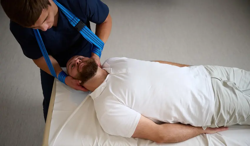 How Much Is Your Whiplash Injury Really Worth? A male chiropractor in a dark blue uniform carefully cradles and manipulates the head and neck of a bearded male patient lying flat on a treatment table in a clinical setting illustrating the specialized chiropractic care required to treat a whiplash injury following a car accident in Georgia.