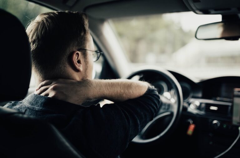A young man with glasses seen from behind in the driver's seat of a car reaching one hand back to grip his neck while the other rests on the steering wheel with blurred scenery visible through the windshield depicting the persistent neck pain and stiffness that drivers commonly experience after suffering a whiplash injury in a rear-end collision and could use legal assistance from Wade Law Office Injury Lawyer.